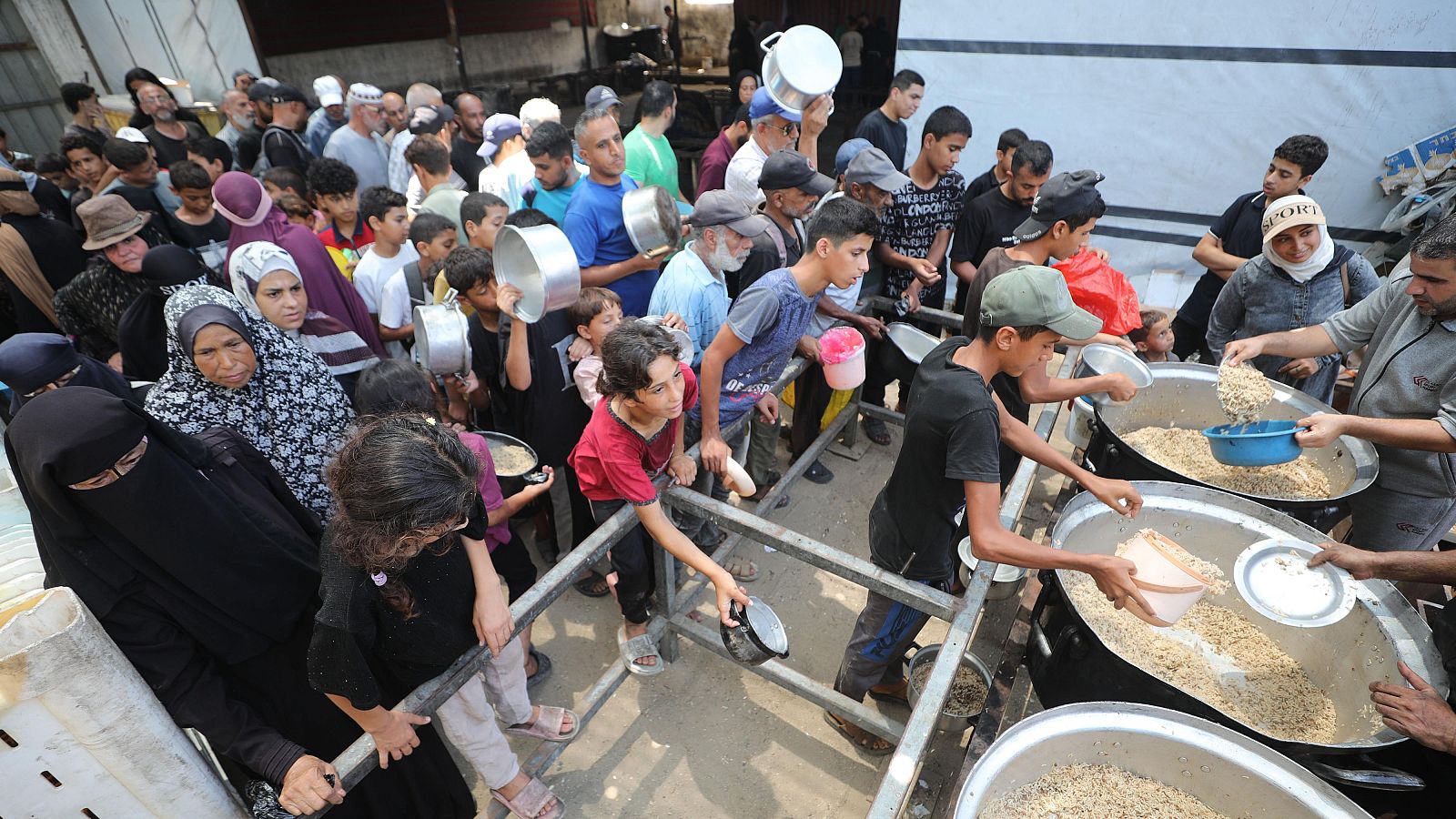 Distribución de alimentos en un campamento, con voluntarios sirviendo arroz a una multitud de personas, incluyendo niños y mujeres con velos.