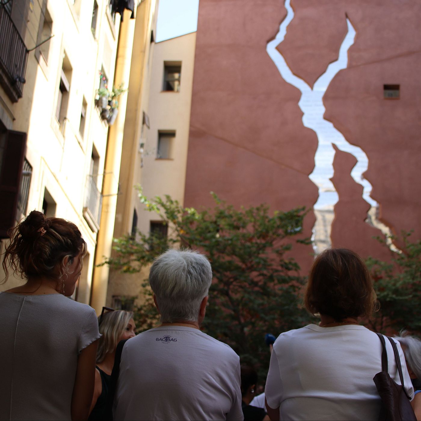 Tres persones contemplen una gran "escletxa" artística en la paret d'un edifici terracota, possiblement simbolitzant un problema social.