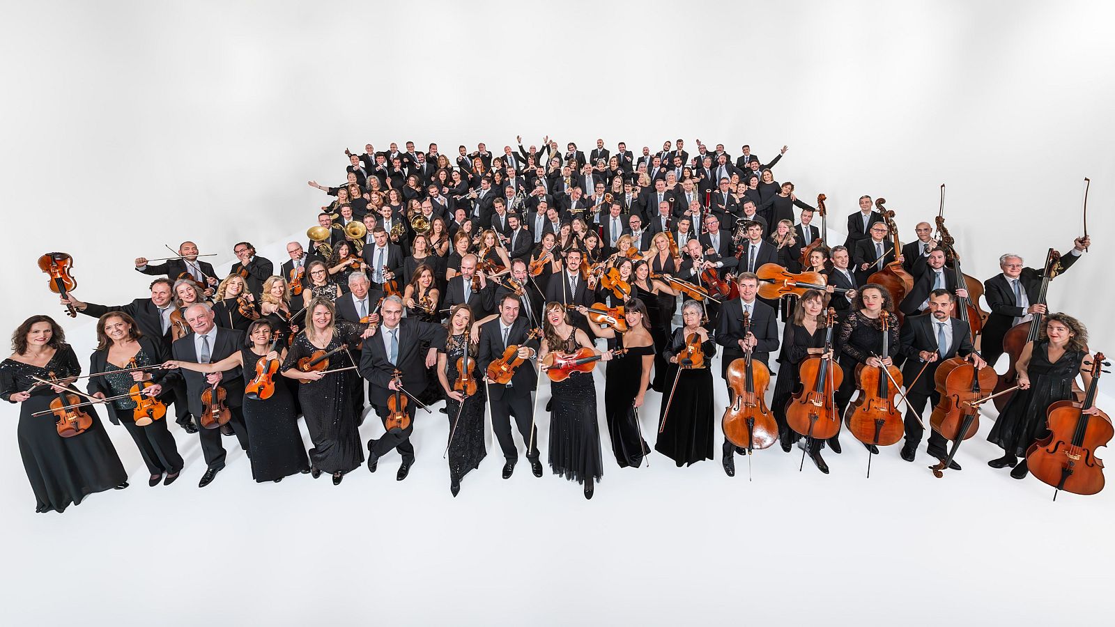Retrato grupal de la Orquesta y Coro de RTVE, con 150-200 miembros en trajes formales, mostrando instrumentos de cuerda, viento-metal y madera sobre fondo blanco.