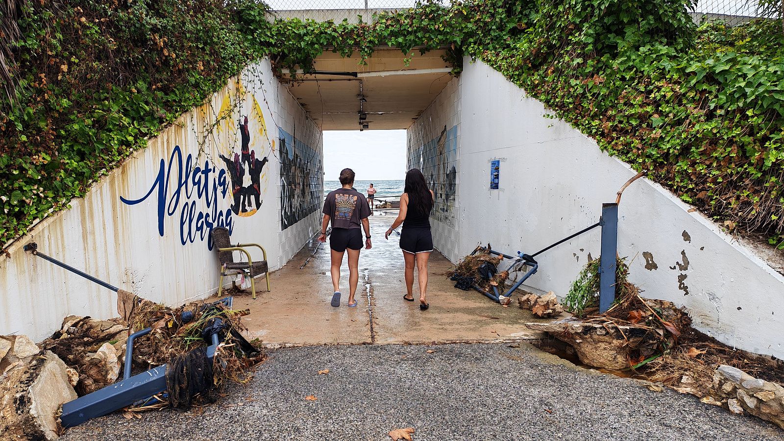 Túnel peatonal cap a "Platja Llarga" amb grafiti, mural de castellers i danys per temporal. Dues persones s'endinsen cap a la sortida al mar.