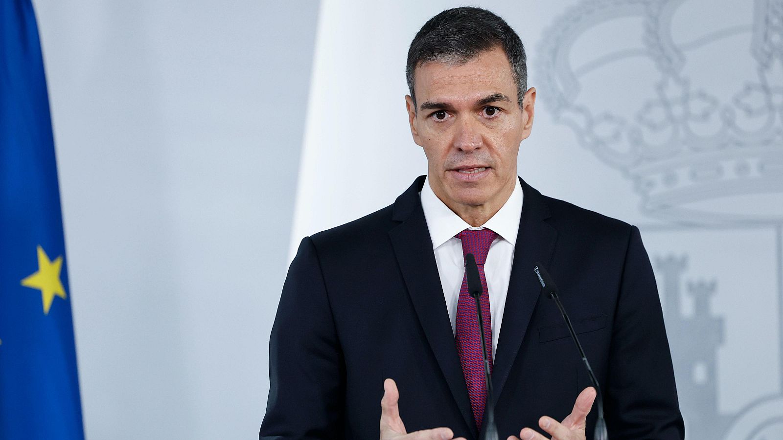 Pedro Sánchez, Presidente del Gobierno de España, en primer plano, con traje oscuro y corbata vino tinto, gesticula mientras habla ante un micrófono. Fondo con banderas de la UE y Escudo de España.
