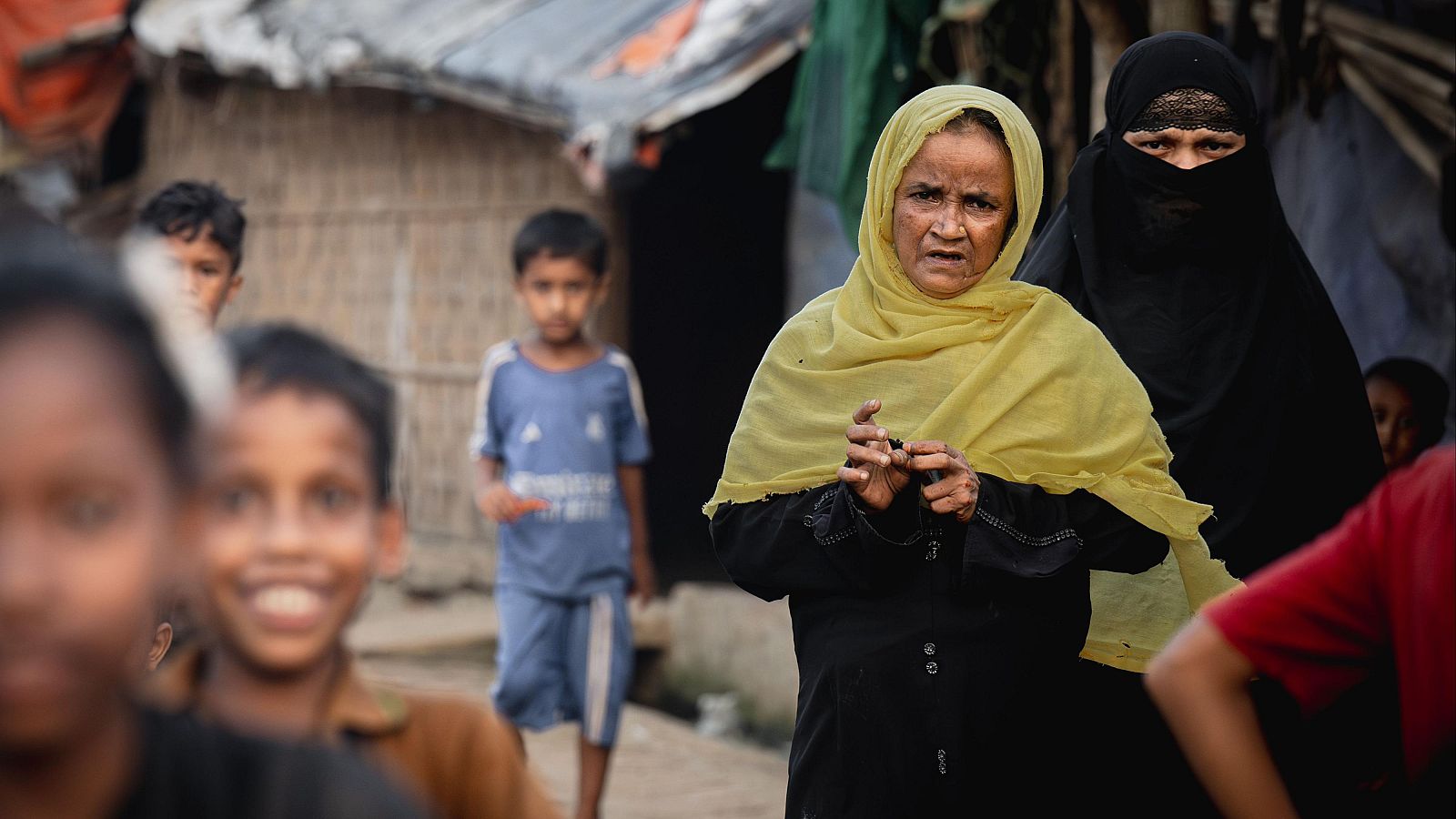 Dos mujeres rohingya, una mayor con hiyab amarillo y otra joven con niqab negro, posan en el campamento de Kutupalong, Bangladés, rodeadas de niños. El fondo revela las humildes construcciones del asentamiento.