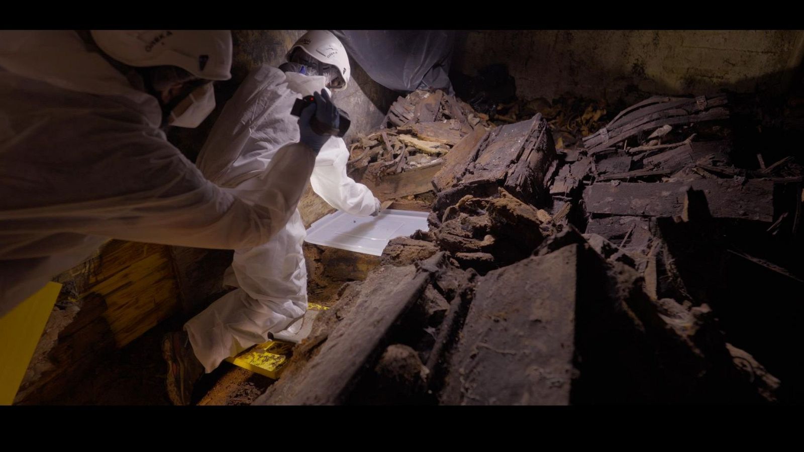Excavación con tierra, huesos humanos desarticulados y restos de madera. Iluminación focalizada resalta texturas. Objeto vertical en el borde derecho.