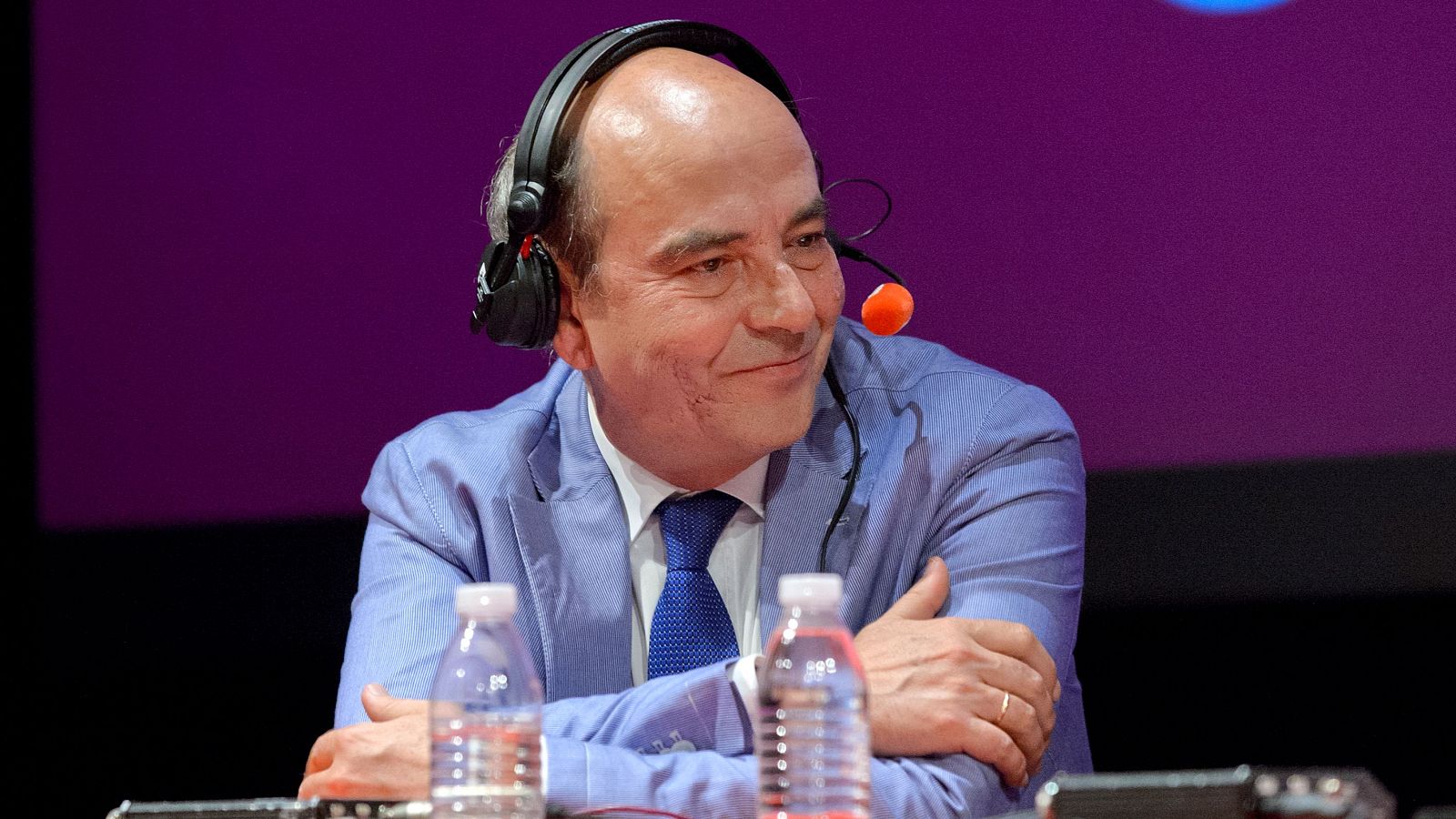 Alfonso Nasarre, hombre calvo de mediana edad, sonríe amablemente con auriculares y micrófono. Viste traje azul claro y camisa blanca. Fondo púrpura.
