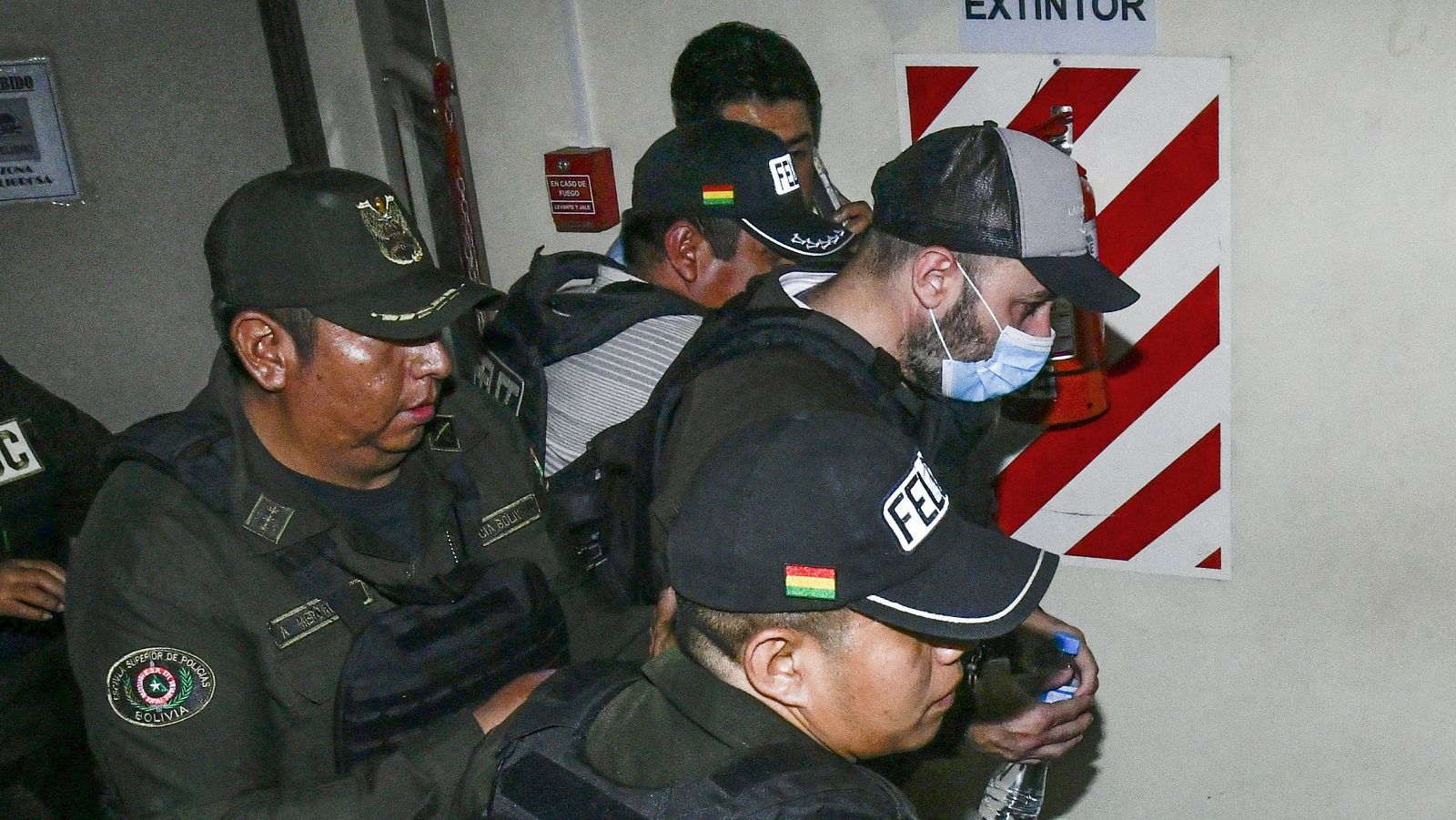 Fernando Cerimedo, empresario argentino, es trasladado detenido por policías bolivianos de la FELCC en un pasillo con señales de extintor y alarma de incendios.