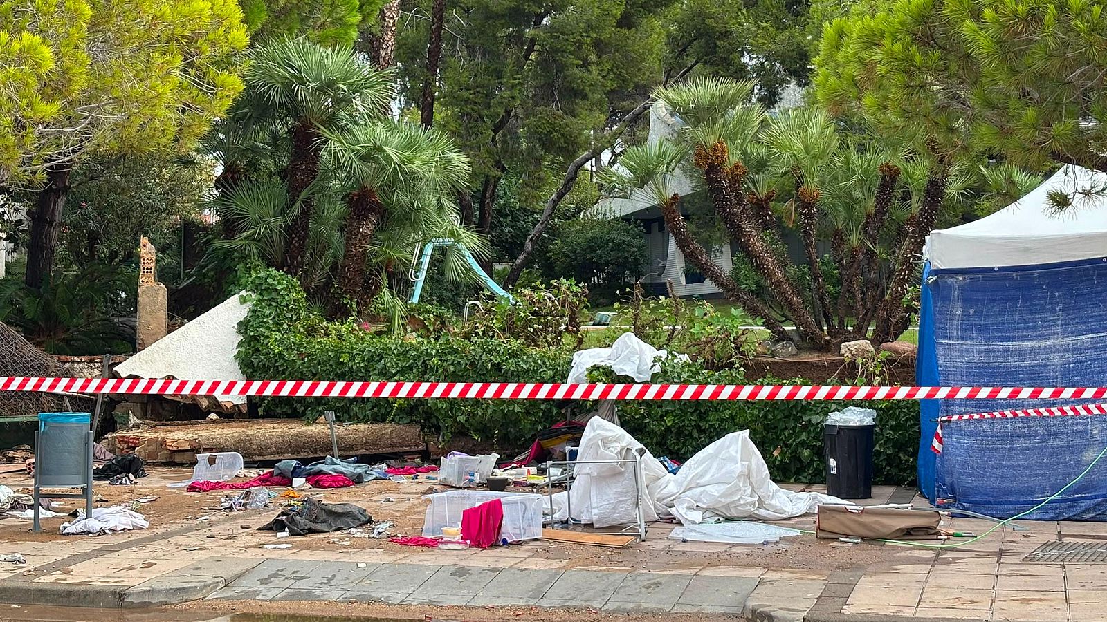 Escena d'un despreniment amb escombres, roba i contenidors a la vorera, separats per cinta de seguretat. Al fons, vegetació, un tobogan i una tenda d'emergència.
