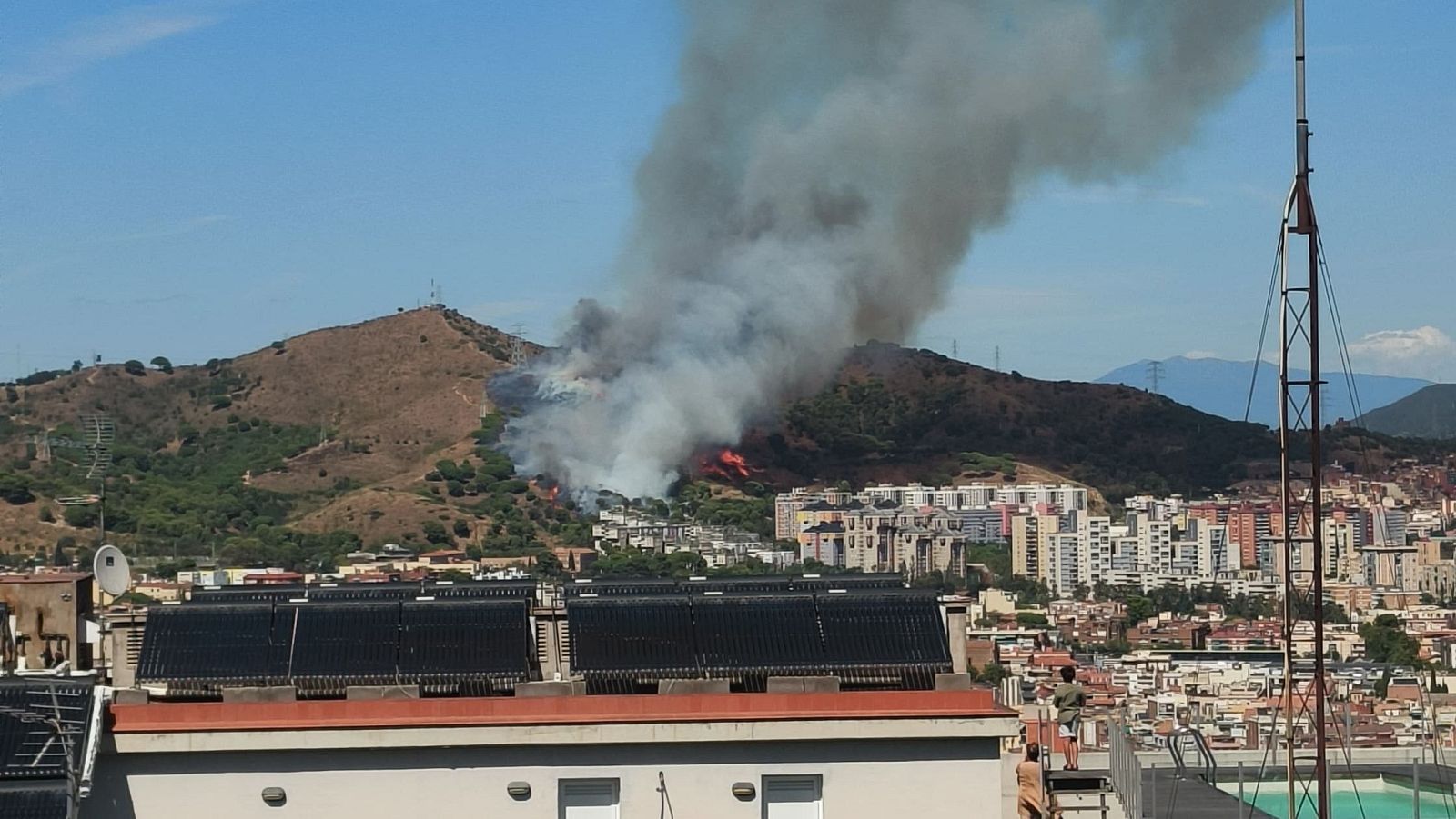 Incendi forestal a la serra de Collserola, amb fum dens sobre una ciutat amb edificis i panells solars. Una antena metàl·lica al primer pla.