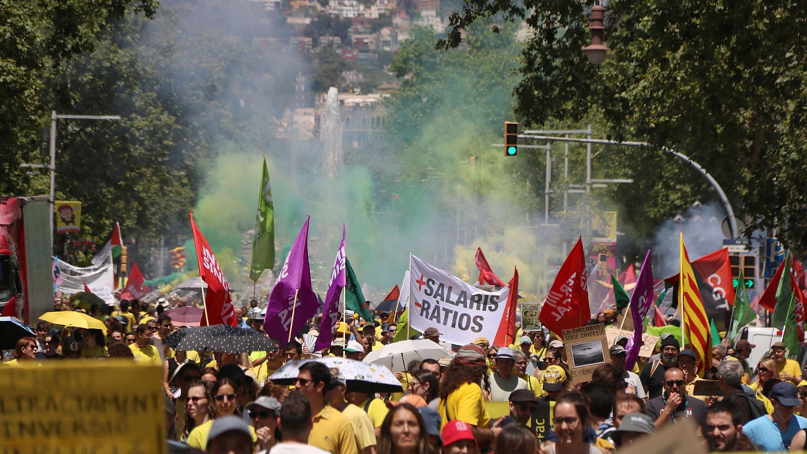 Manifestació de docents a Barcelona amb samarretes grogues, pancartes reivindicatives i banderes diverses. El sol brilla i s'observen para-sols.