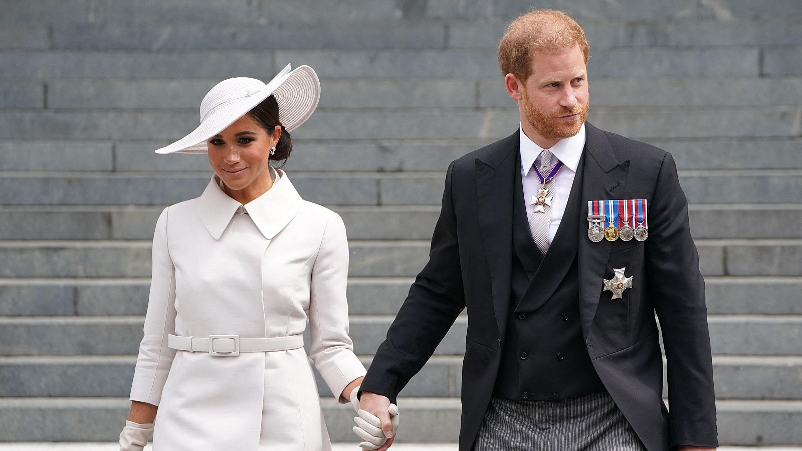 Príncipe Harry y Meghan Markle, Duques de Sussex, tomados de la mano, posan ante una imponente escalera de piedra. Ella viste de blanco roto, él de chaqué oscuro con condecoraciones.
