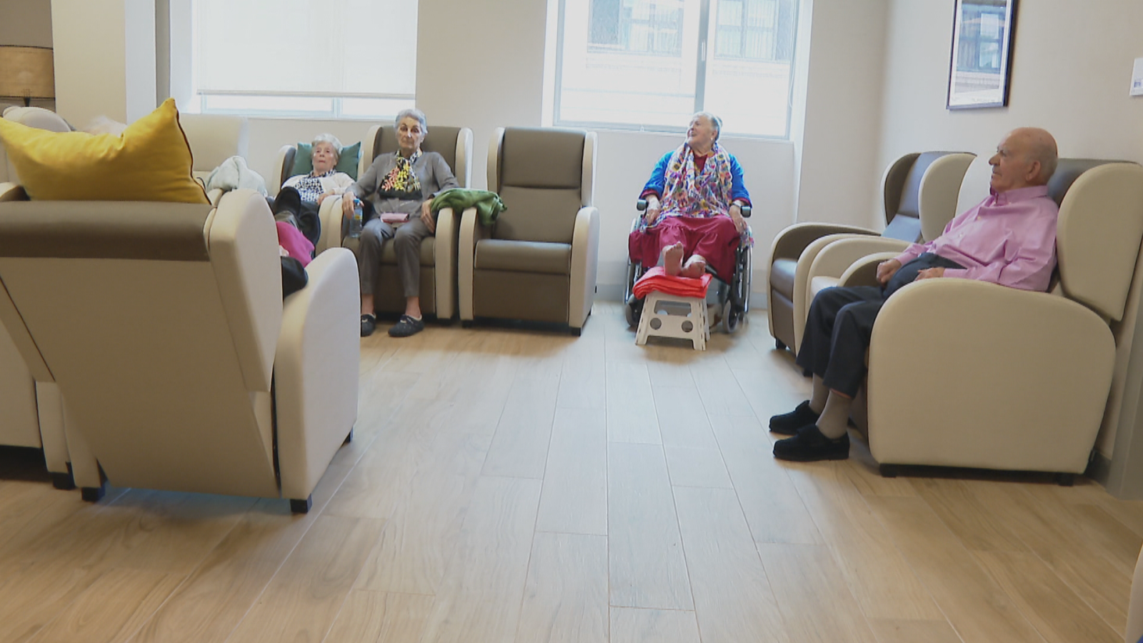 Cinco personas mayores descansan en sillones de una sala de estar moderna y luminosa, con grandes ventanales y suelo de parquet. Una mujer en silla de ruedas observa atentamente.