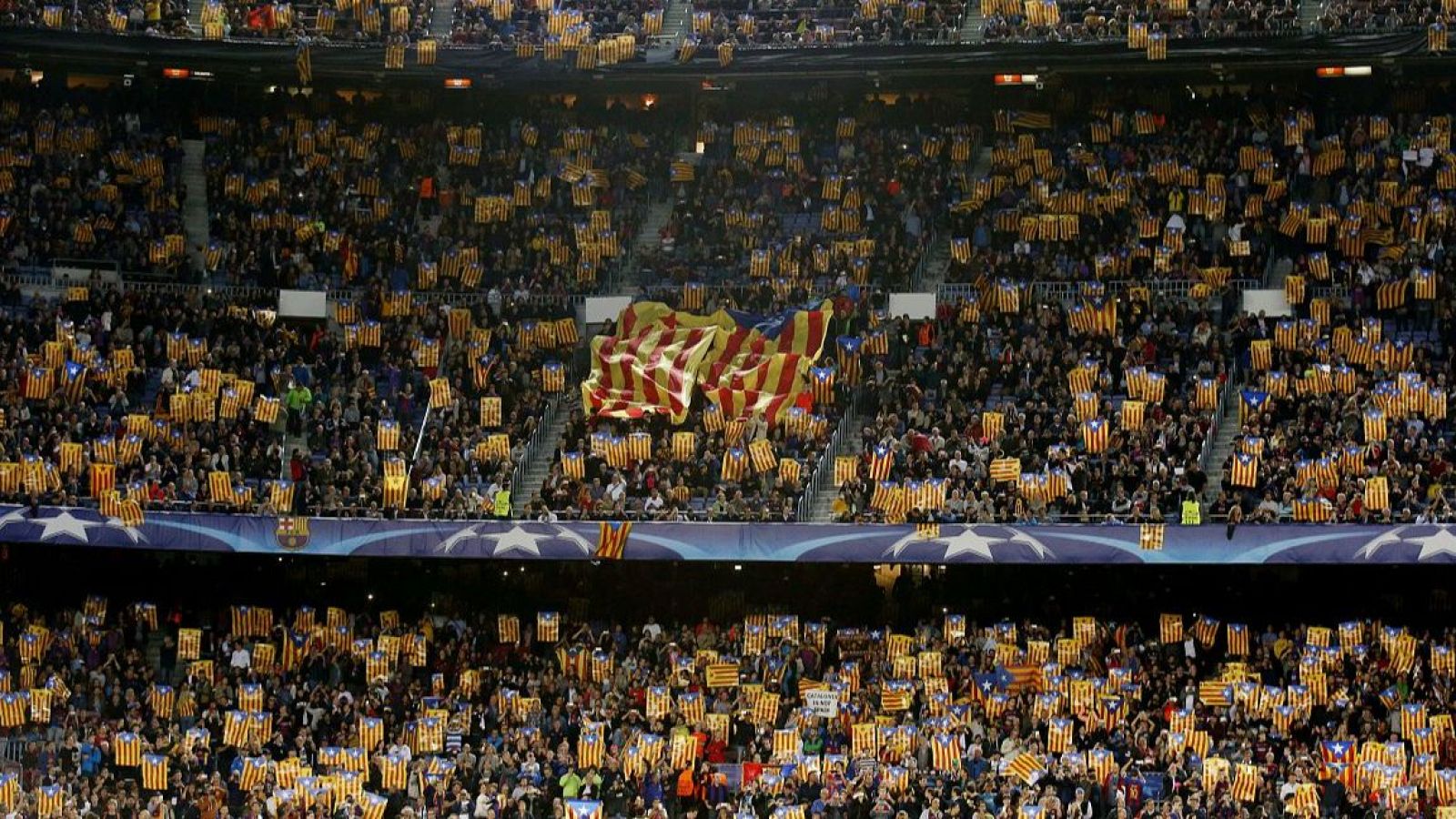 Vista panorámica de un estadio de fútbol nocturno repleto de aficionados ondeando banderas catalanas (Senyera y Estelada) y un banner de la UEFA Champions League.
