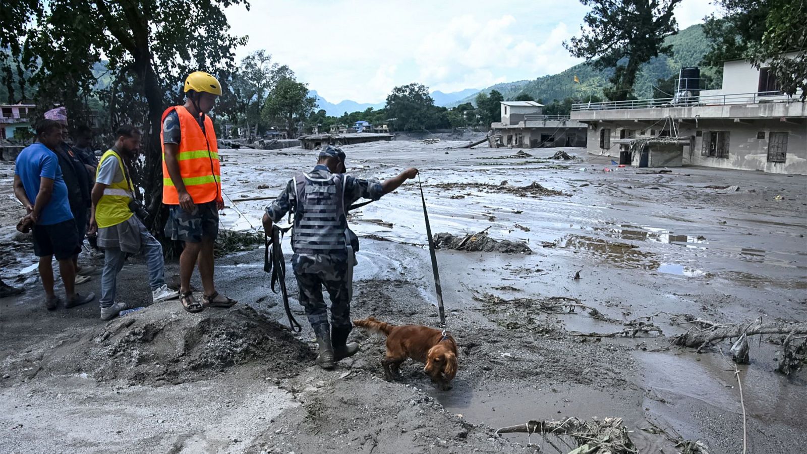 Un hombre uniformado y su perro de búsqueda inspeccionan una zona devastada por una riada en Nepal, con otros observadores y edificios dañados al fondo.