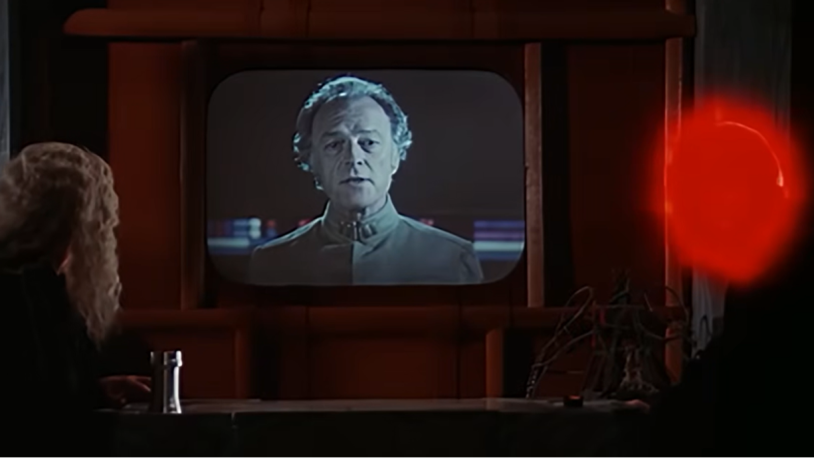 Bernard Behrens como Comandante en pantalla retro, con persona de cabello claro observando. Luz roja intensa a la derecha.