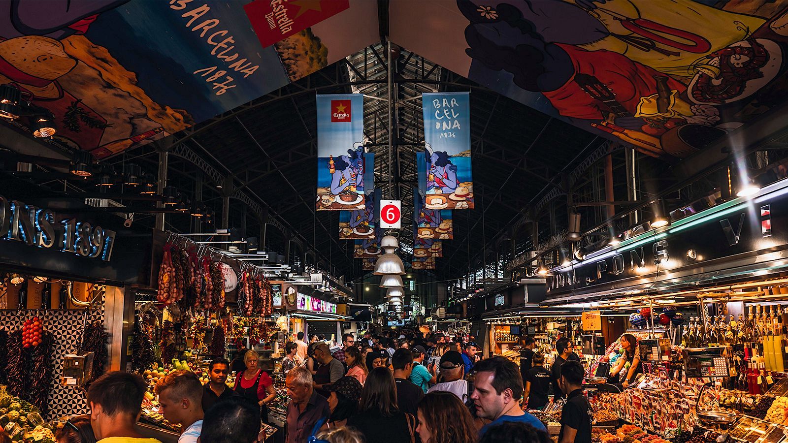 En el interior de un mercado, se aprecian numerosos puestos con productos frescos y elaborados, como frutas, verduras, carnes y embutidos. Vendedores y compradores interactúan en un ambiente bullicioso, bajo una estructura que soporta publicidad.