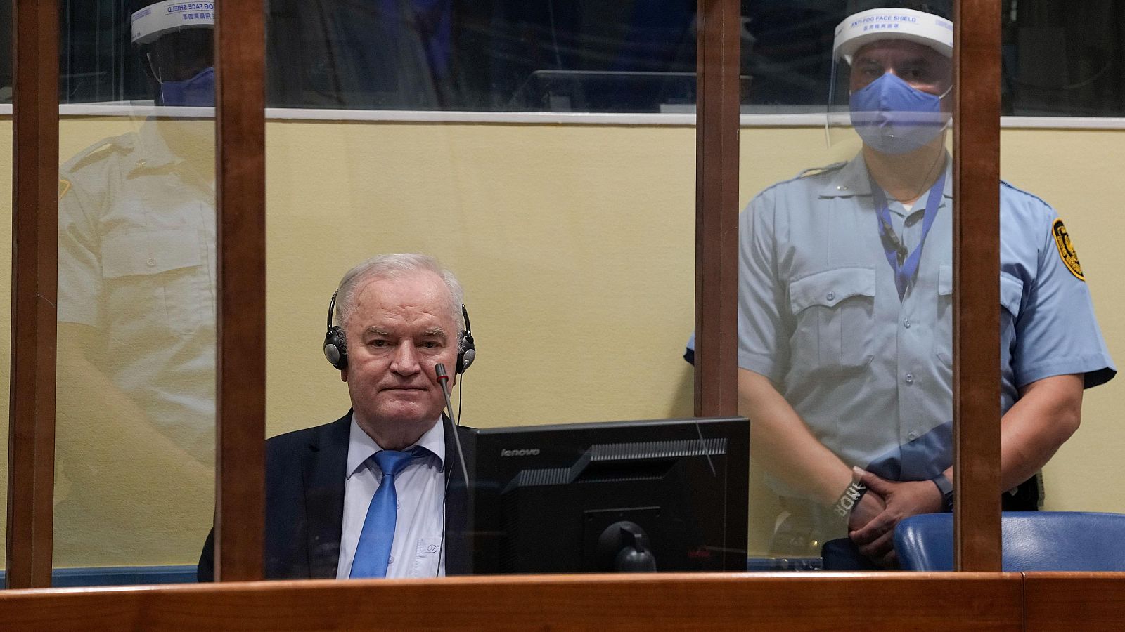 Ratko Mladić, exjefe militar serbobosnio, en una sala de tribunal en La Haya, con auriculares y traje oscuro. Guardias de seguridad visibles tras una mampara de cristal.