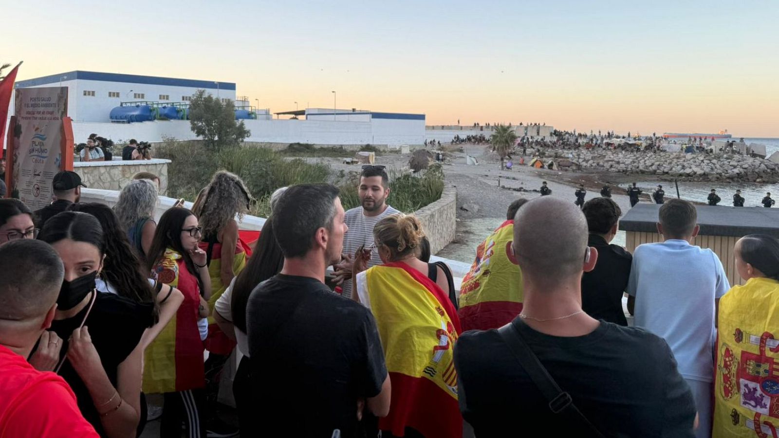 Manifestantes con banderas de España en la Playa Benítez de Ceuta, con presencia de fuerzas de seguridad y una multitud en la arena.