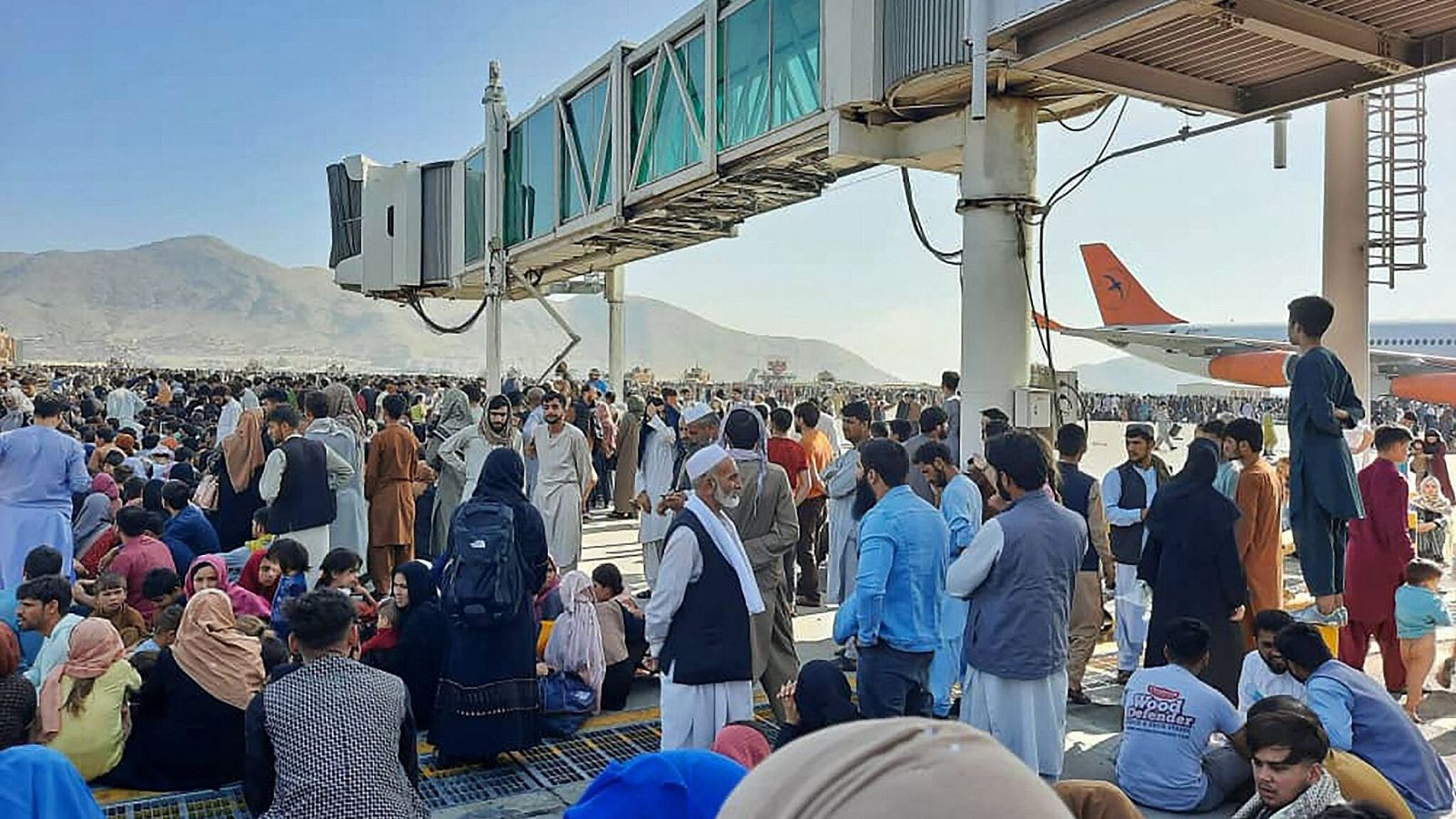 Miles de personas, vestidas con ropas tradicionales de Oriente Medio o Asia del Sur, abarrotan la pista de un aeropuerto, con una pasarela de embarque y la cola naranja de un avión al fondo.