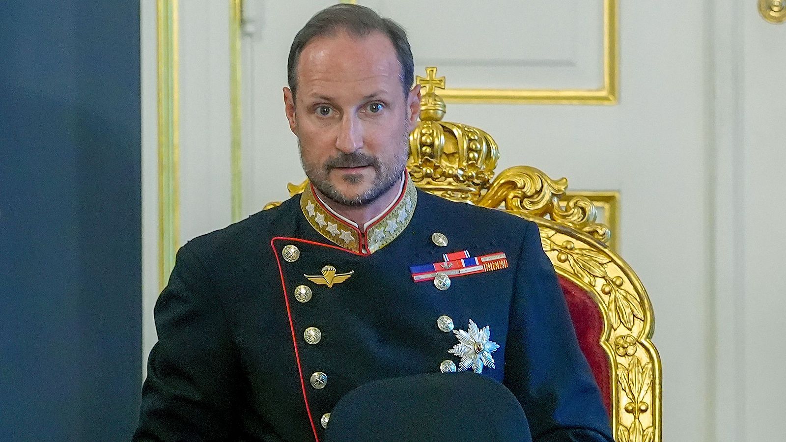 Primer plano del Príncipe Heredero Haakon de Noruega con uniforme militar de gala azul oscuro, condecoraciones y barba, sentado en una silla dorada ornamentada.