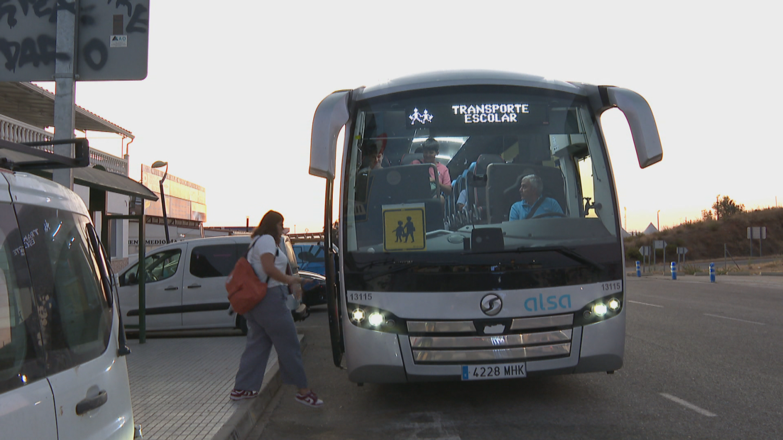 Autobús de "TRANSPORTE ESCOLAR" de Alsa (matrícula 4228 MHK, serie 13115) recogiendo pasajeros. Mujer sube, conductor y niños dentro. Entorno urbano con carretera y edificios. Luz cálida.