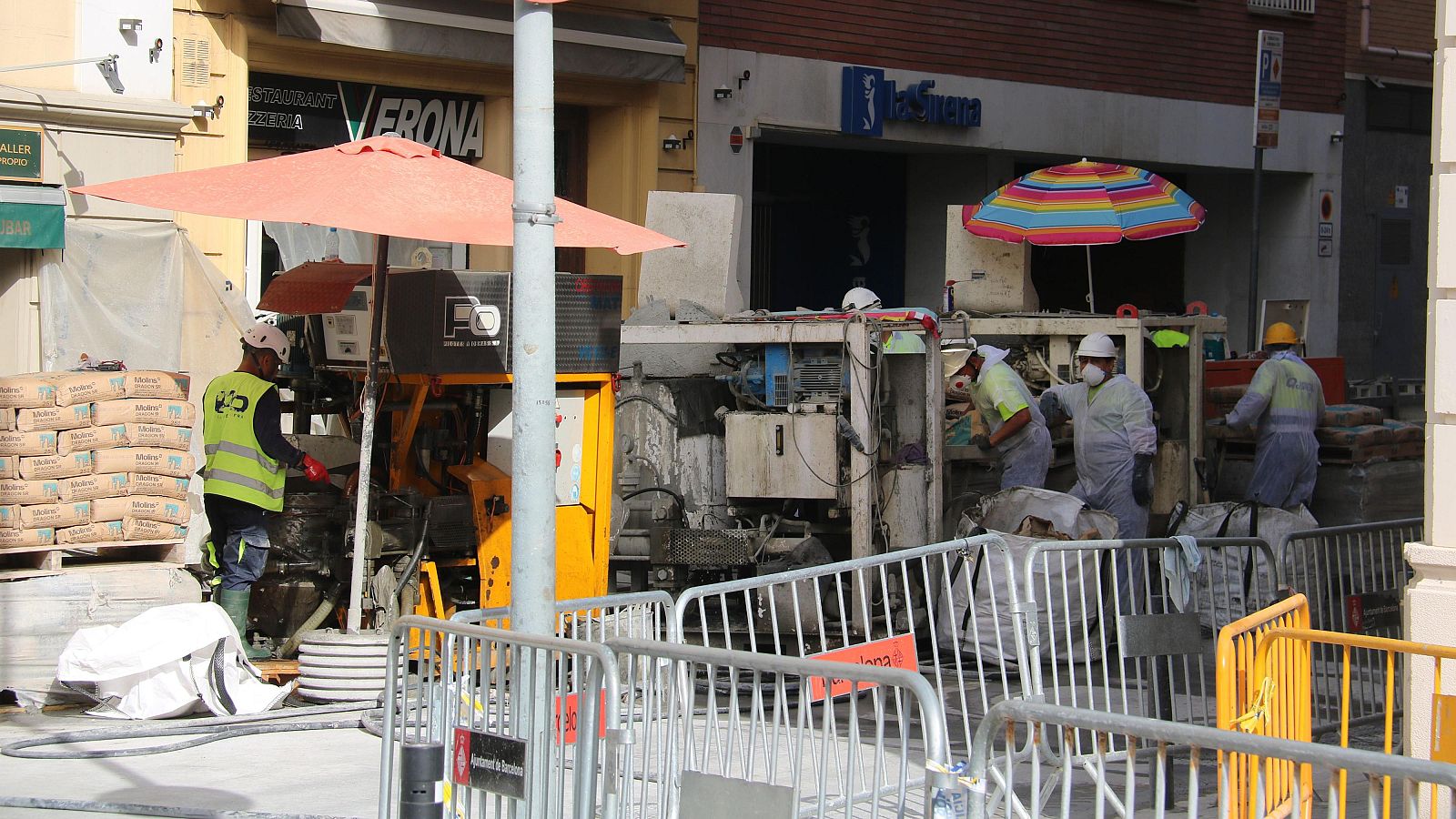 Operarios trabajan en una obra urbana con maquinaria, sacos de cemento y vallas de seguridad. Se aprecian edificios con letreros de "RESTAURANT VERONA" y "la Caixa".