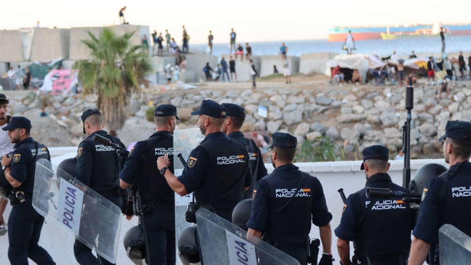 Policías nacionales con escudos y uniformes vigilan una zona costera en Ceuta, donde una multitud se congrega cerca de un muro y refugios improvisados.