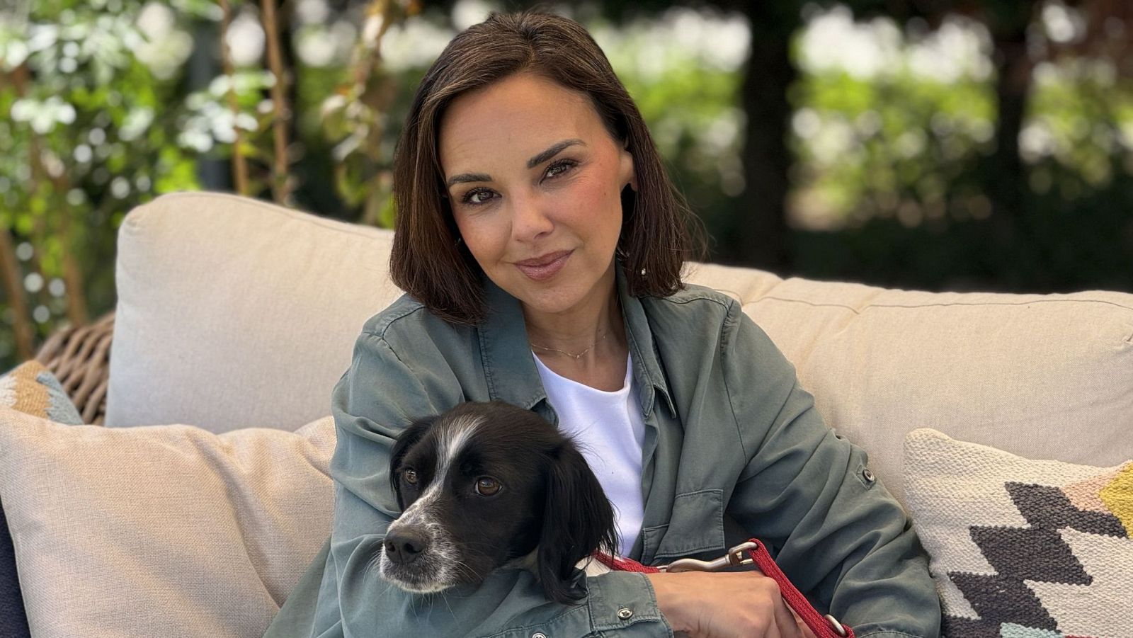 Anne Igartiburu, presentadora española, sonríe a cámara mientras abraza a un perro blanco y negro en un sofá de exterior rodeado de vegetación.