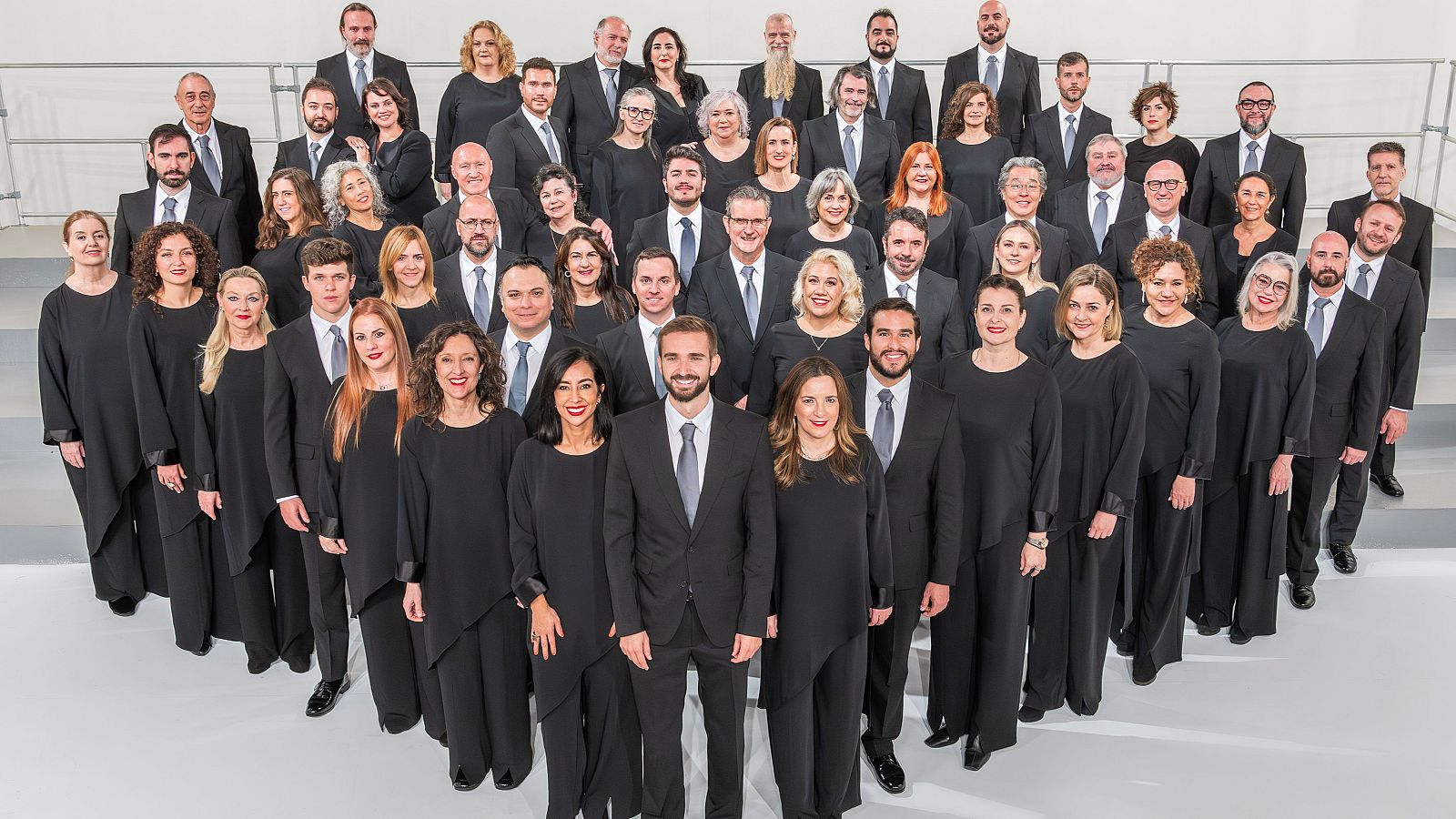 Retrato grupal de 50-60 personas, hombres y mujeres, en atuendo formal oscuro, dispuestos en formación de "V" o semicírculo, mirando a la cámara.