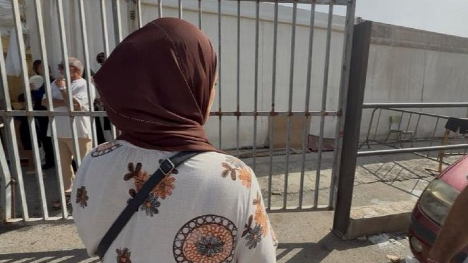 Mujer con hiyab y blusa estampada de espaldas a una verja metálica, con un campamento y un coche rojo al fondo.