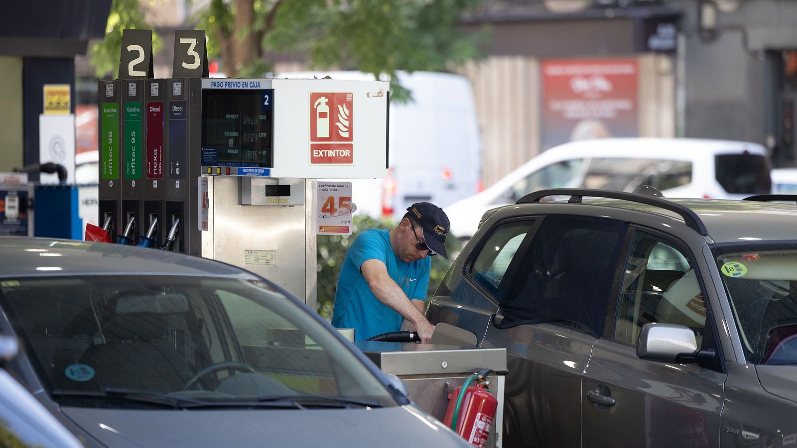 Hombre repostando SUV gris en gasolinera. Surtidores con combustibles y pago previo. Extintor rojo visible.
