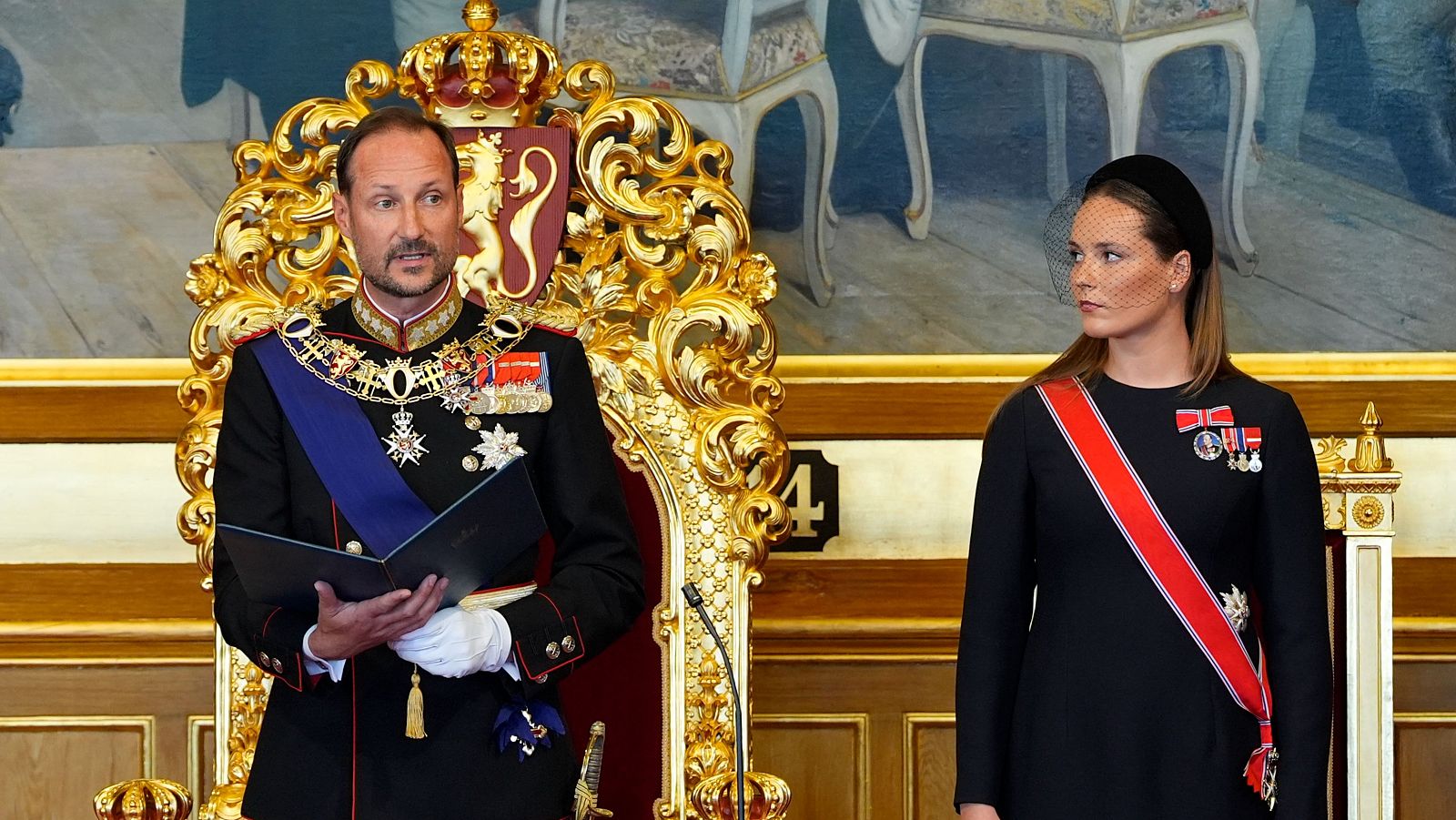 Rey Haakon VIII de Noruega en trono dorado, con uniforme militar y condecoraciones, sostiene un documento. A su derecha, una mujer con diadema y velo observa.