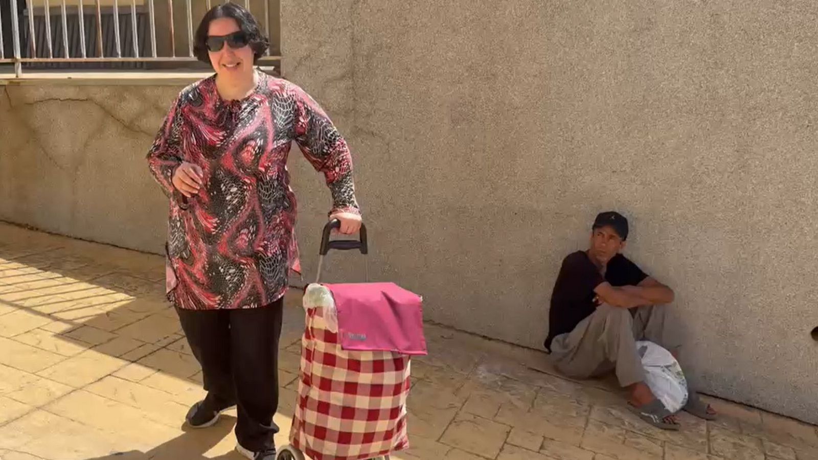 Mujer con carro de la compra y hombre sentado en el suelo, ambos en un espacio exterior pavimentado junto a una pared clara.