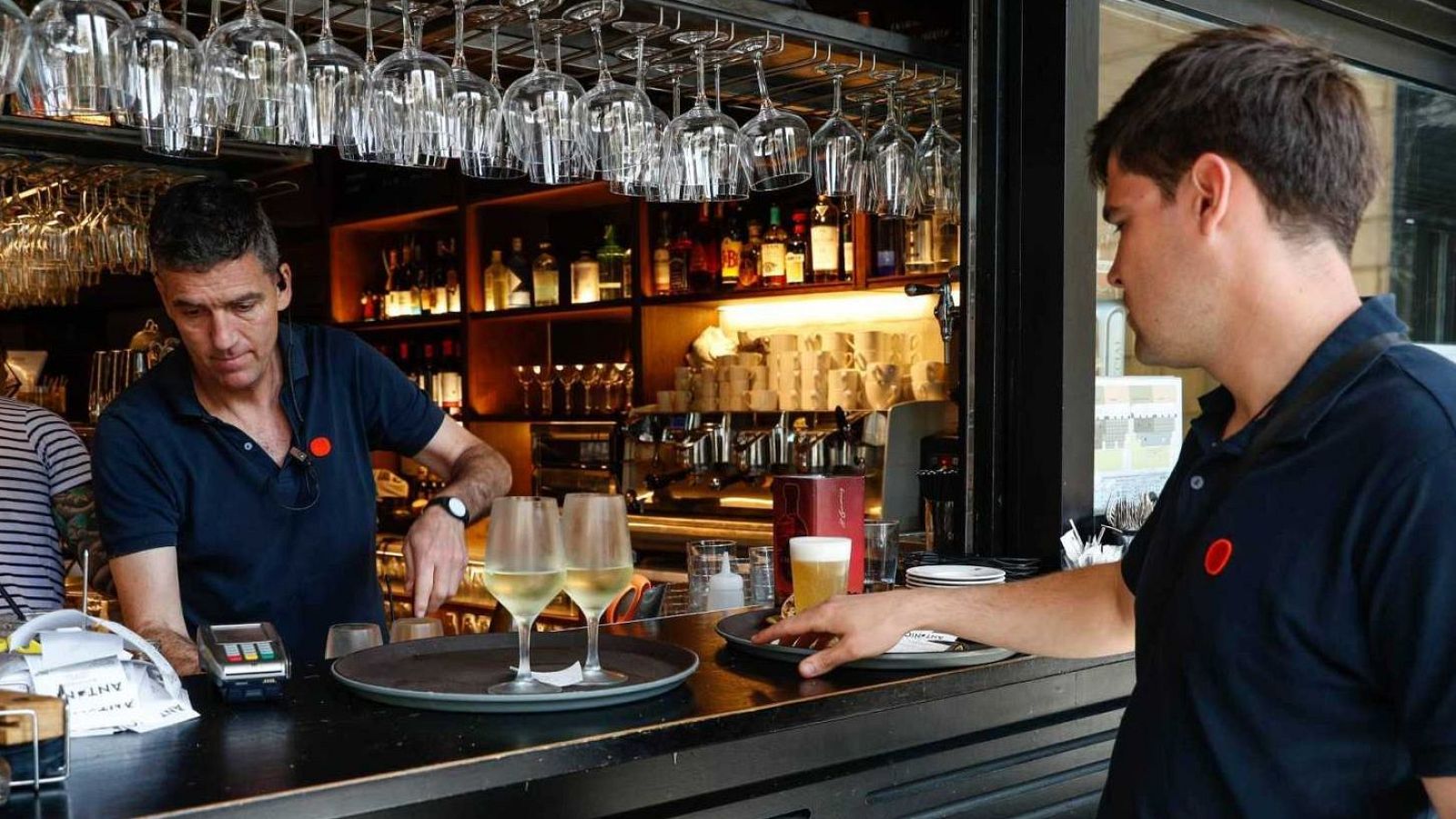 Cambrer amb polo blau i pin vermell serveix cervesa a un client. Detalls del bar: copes penjades, ampolles, TPV.
