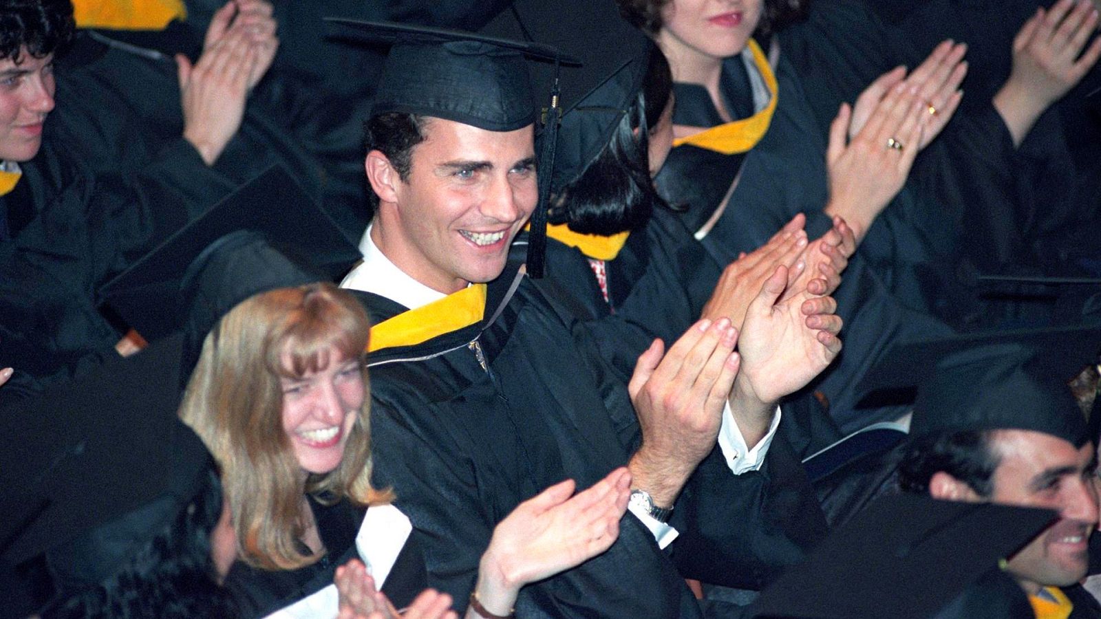 Joven rey Felipe VI, con toga y birrete amarillos, aplaude en una ceremonia de graduación universitaria. Ambiente festivo y solemne.