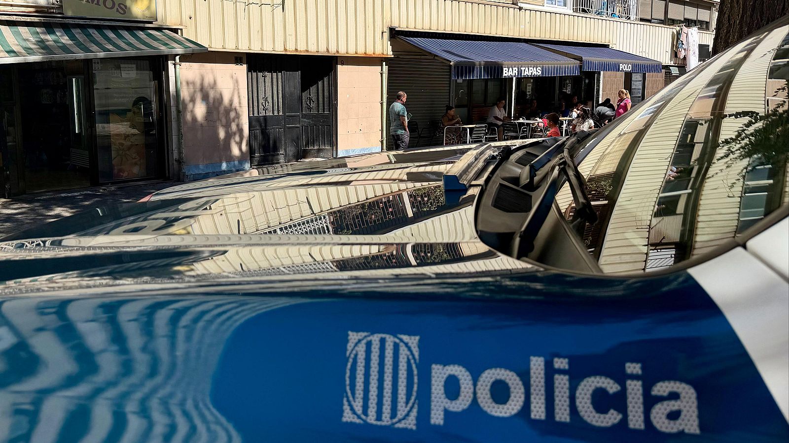 Detalle de un coche policial de los Mossos d'Esquadra con "policia" y emblema, reflejando edificios y un bar con terraza al otro lado de la calle.