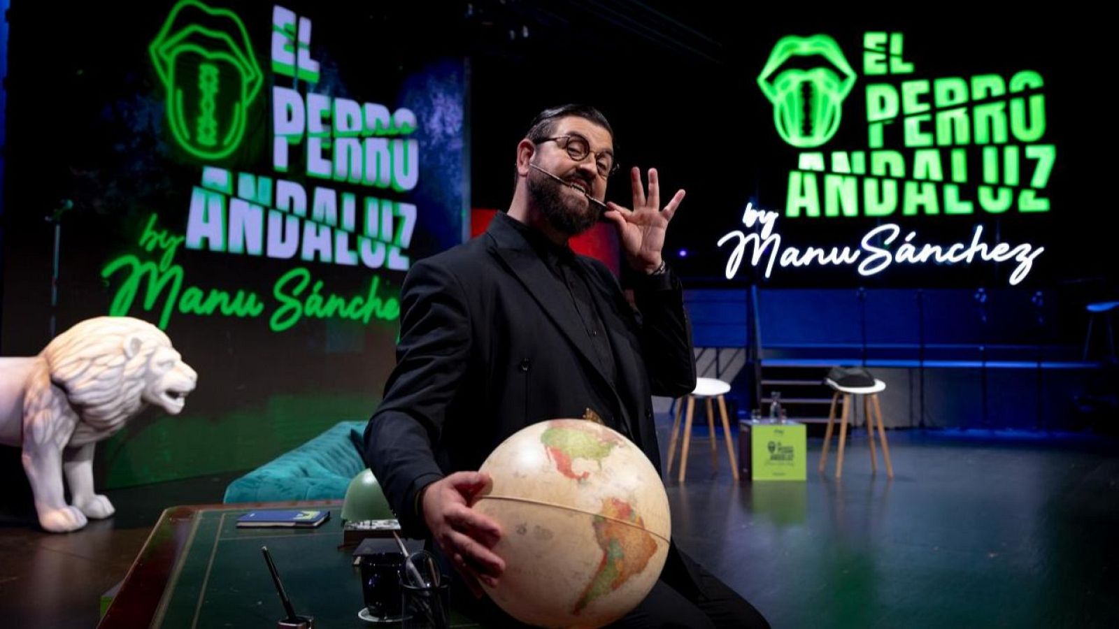 Manu Sánchez, con gafas y traje, presenta "EL PERRO ANDALUZ" en un escenario de neón verde, sosteniendo un globo terráqueo y con un objeto en la boca. La pantalla muestra el título y un logo.