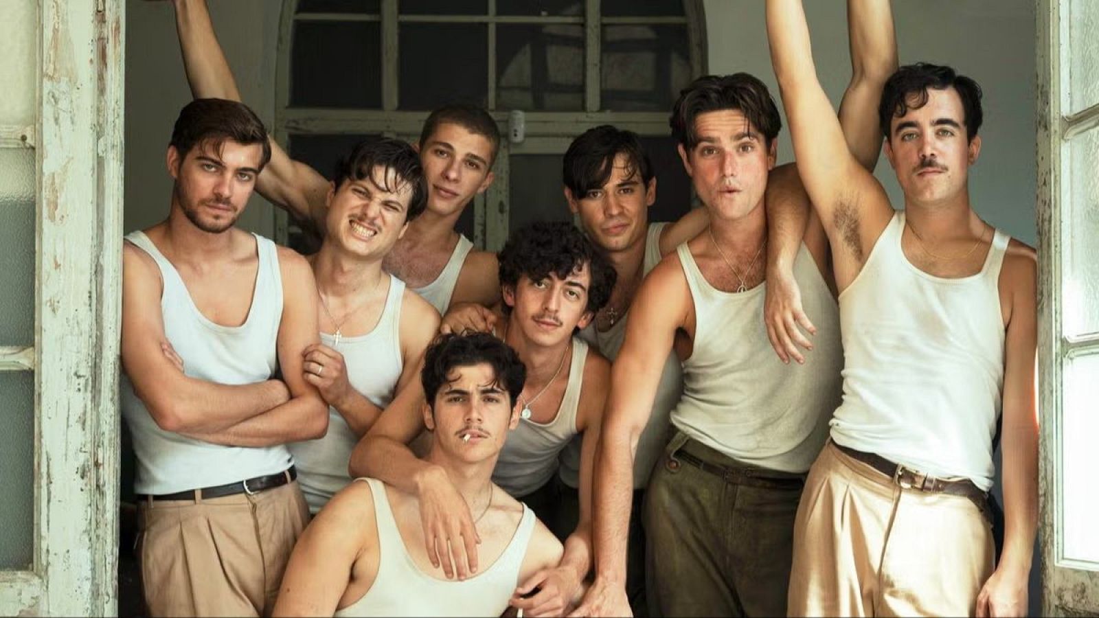Grupo de ocho hombres con camisetas blancas y pantalones de colores posan en un marco de puerta deteriorado, evocando una estética de los años 70.