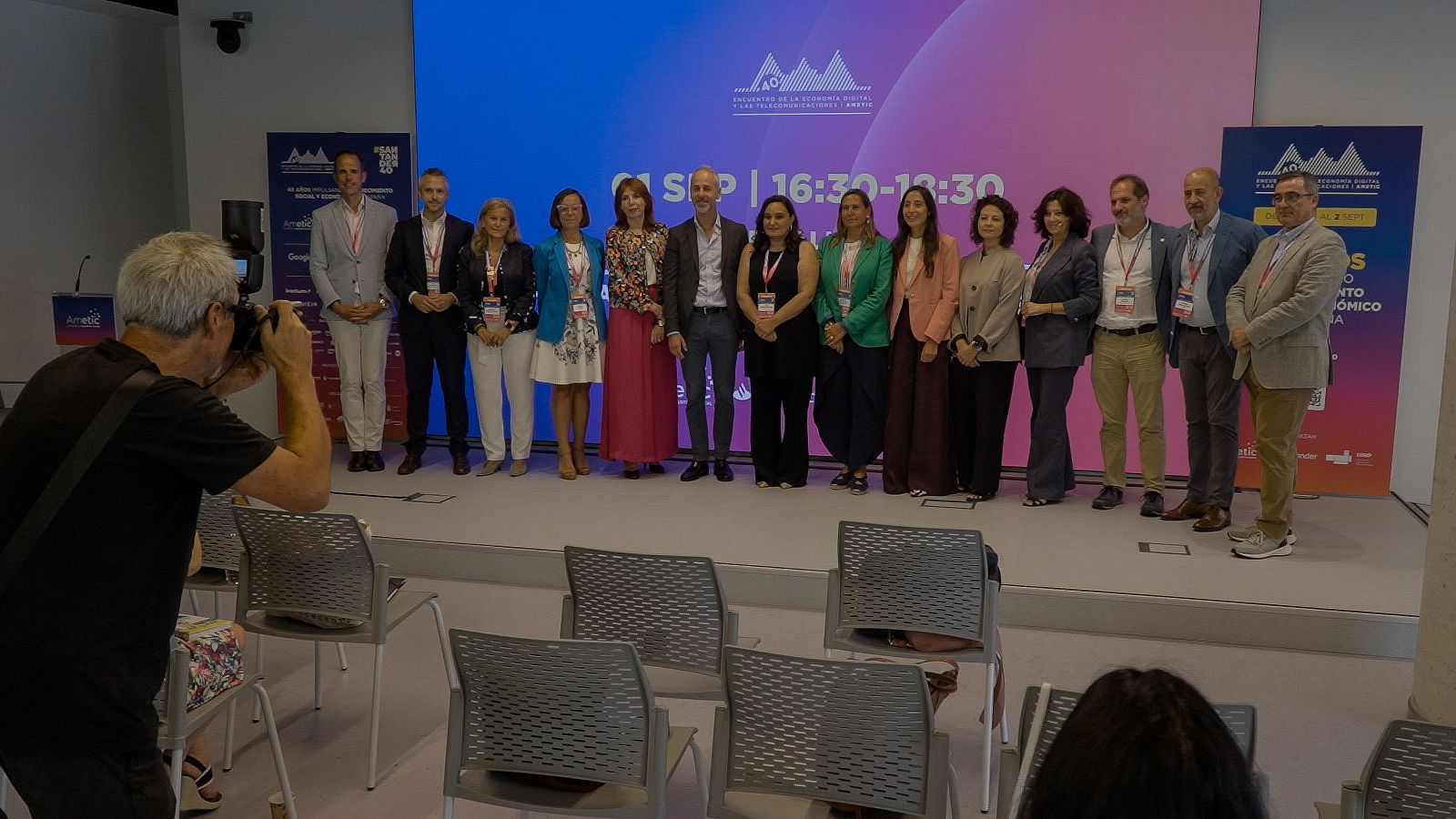 Grupo de catorce personas en un escenario con pantalla que muestra "UNIVERSIDAD DE LA ECONOMÍA DIGITAL Y LAS TRANSFORMACIONES" y "1 SEP | 16:30-18:30".