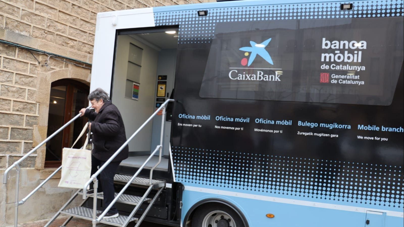 Mujer mayor subiendo escaleras a oficina bancaria móvil de CaixaBank. Vehículo con logotipos de CaixaBank y Generalitat de Catalunya.