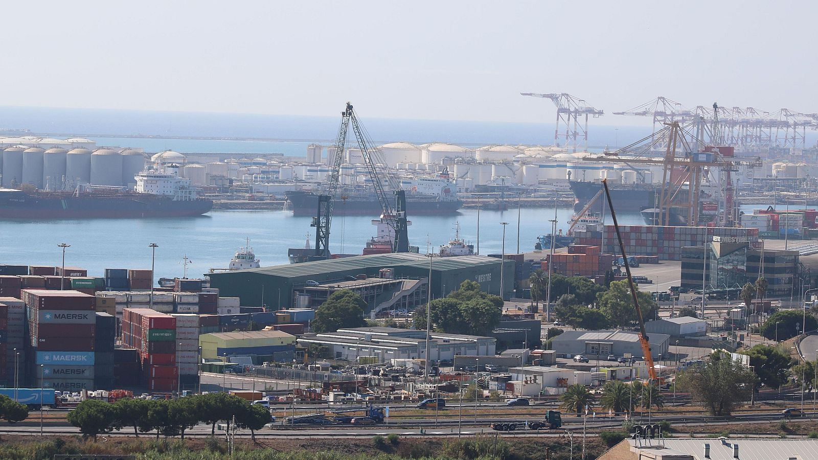 Vista del Puerto de Barcelona con buques, grúas, contenedores (algunos con la marca MAERSK), vehículos y tanques de almacenamiento en el horizonte.