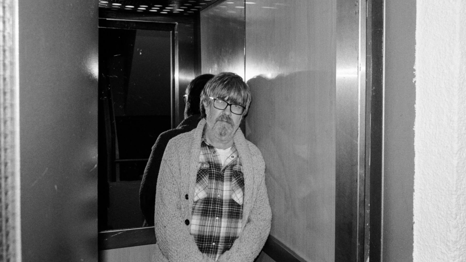 Santiago Segura, con barba canosa y gafas, posa pensativo en un ascensor con camisa de cuadros y rebeca. Reflejo en pared metálica.