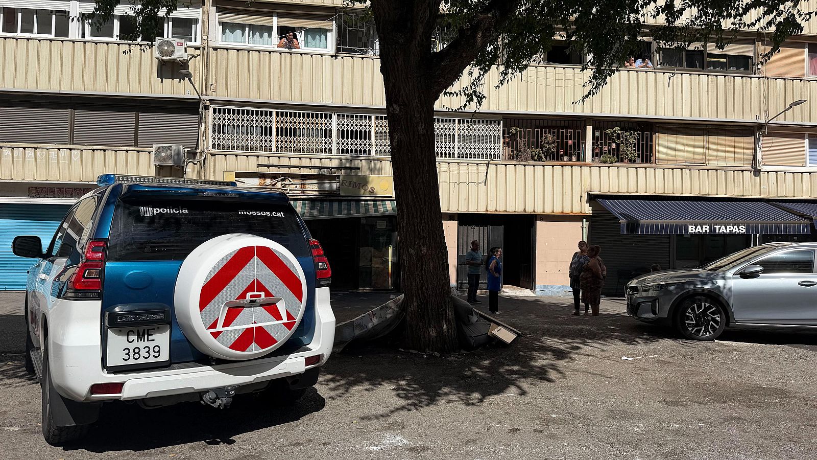 Vehicle policial dels Mossos d'Esquadra davant d'un edifici amb un bar. Es veuen persones a la vorera i als balcons.