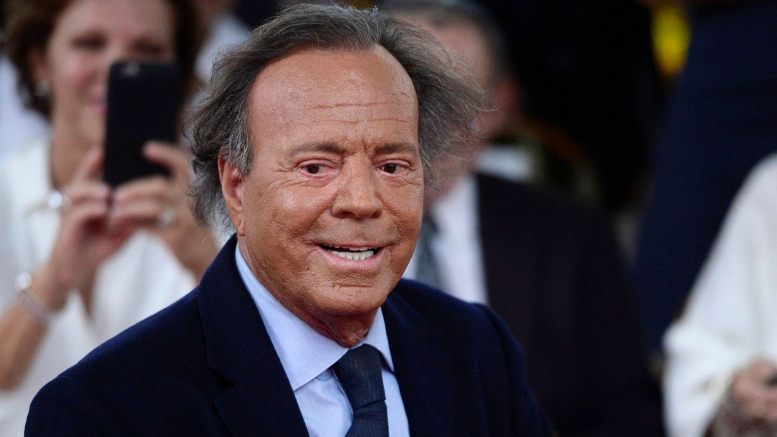 Julio Iglesias posa sonriente, vestido con un traje azul oscuro, camisa celeste y corbata a juego, mirando a la cámara. Se aprecia su cabello castaño con canas.