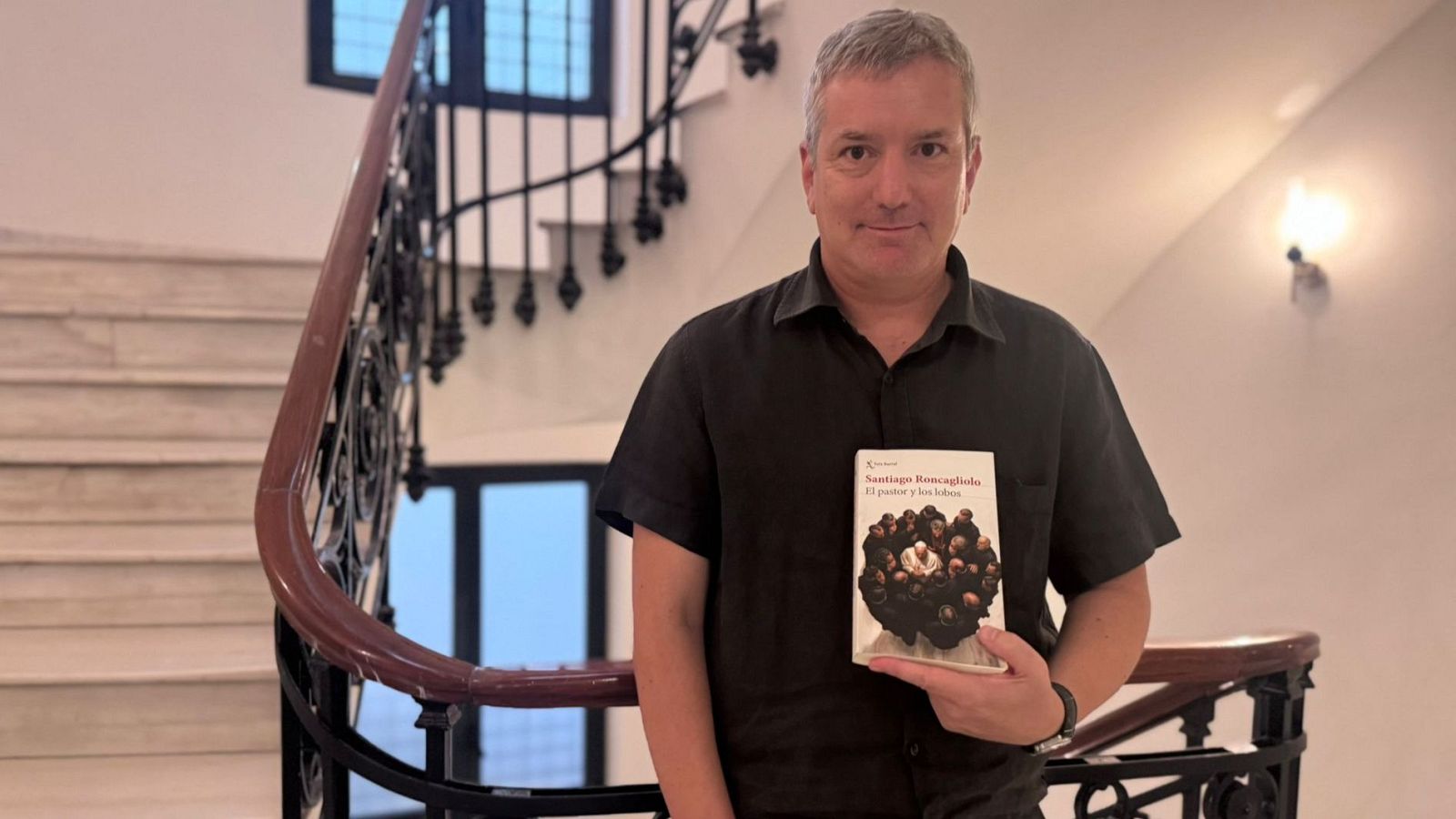 Santiago Roncagliolo, autor de "El pastor y los lobos", sonríe a la cámara sosteniendo su libro en un interior con una escalera ornamentada.