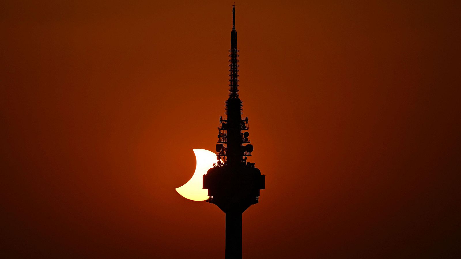 Silueta de torre de telecomunicaciones con antenas contra un cielo cálido y un sol eclipsado en forma de media luna.