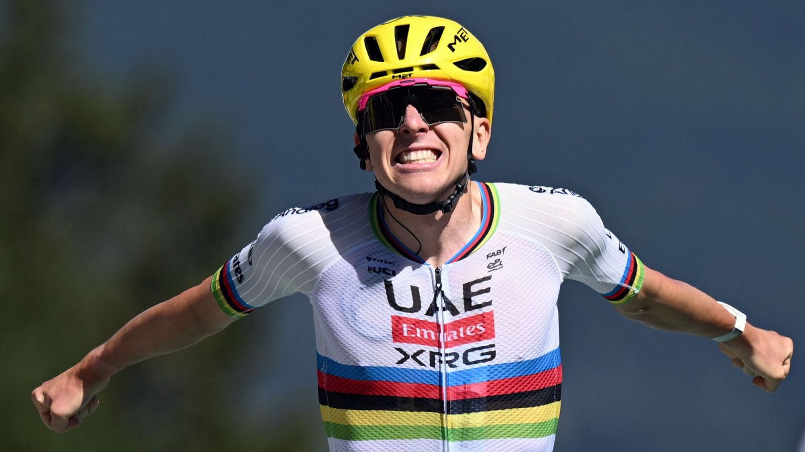 Tadej Pogacar celebra con euforia, luciendo casco MET amarillo, gafas oscuras con borde rosa y maillot blanco de UAE Emirates con franjas arcoíris.