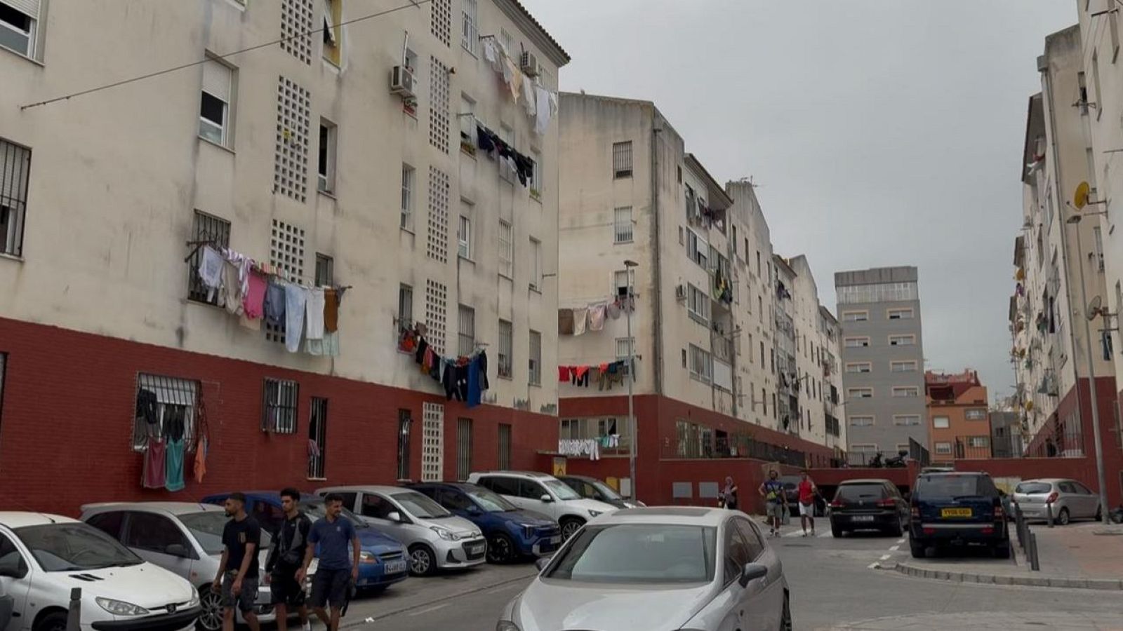 Vista de una calle residencial en Ceuta, con edificios de dos tonos, ropa tendida y coches aparcados. Tres hombres jóvenes transitan por la acera en primer plano.