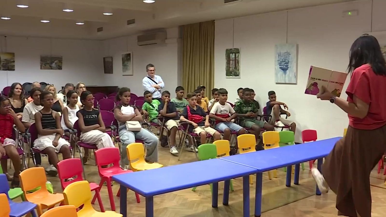 Niños saharauis escuchan atentamente a una cuentacuentos en una sala luminosa con sillas de colores. La cuentacuentos, de espaldas, muestra un libro ilustrado.