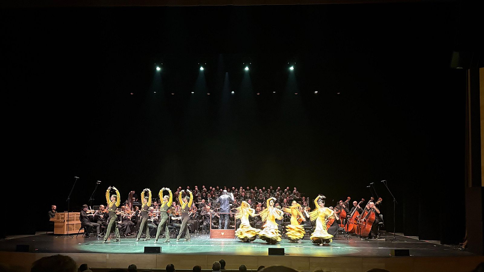 Espectáculo en vivo con bailarines en tops amarillos y vestidos flamencos dorados, orquesta sinfónica y coro, bajo focos intensos en un gran escenario.