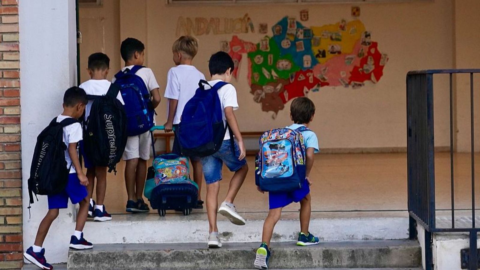 Niños varones con mochilas, algunos con ruedas, suben escaleras hacia una escuela. Al fondo, un mapa de Andalucía con el texto "ANDALUCÍA".