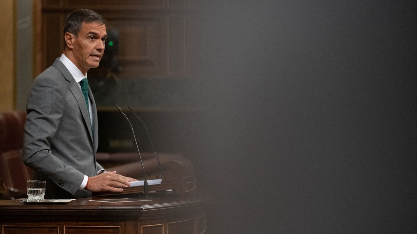 Pedro Sánchez, Presidente del Gobierno, habla en un atril durante una sesión plenaria en el Congreso de los Diputados, vestido con traje gris y corbata verde esmeralda.
